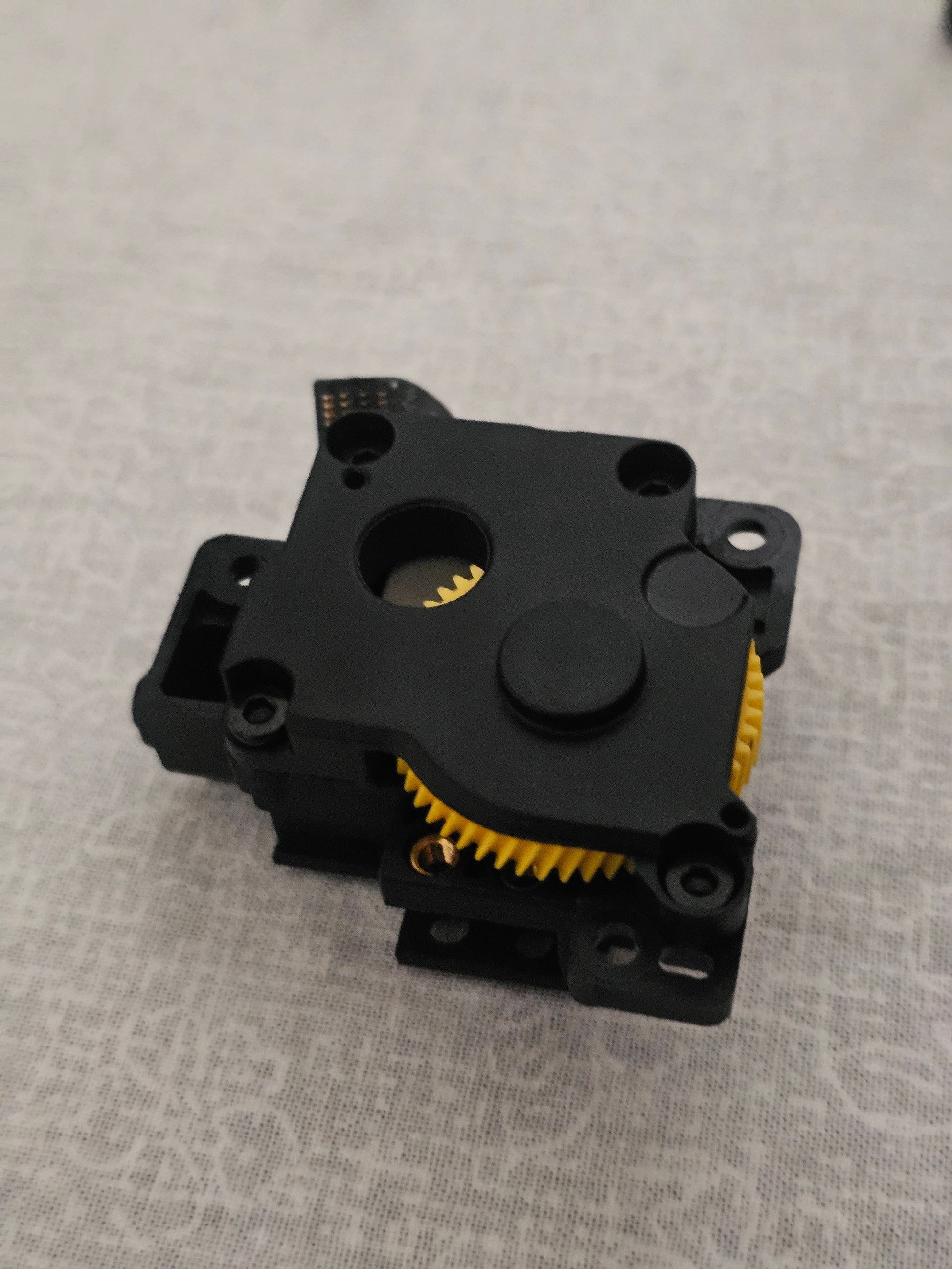 A black plastic gear motor or encoder component with yellow teeth visible on the side, photographed on a light gray textured surface.
