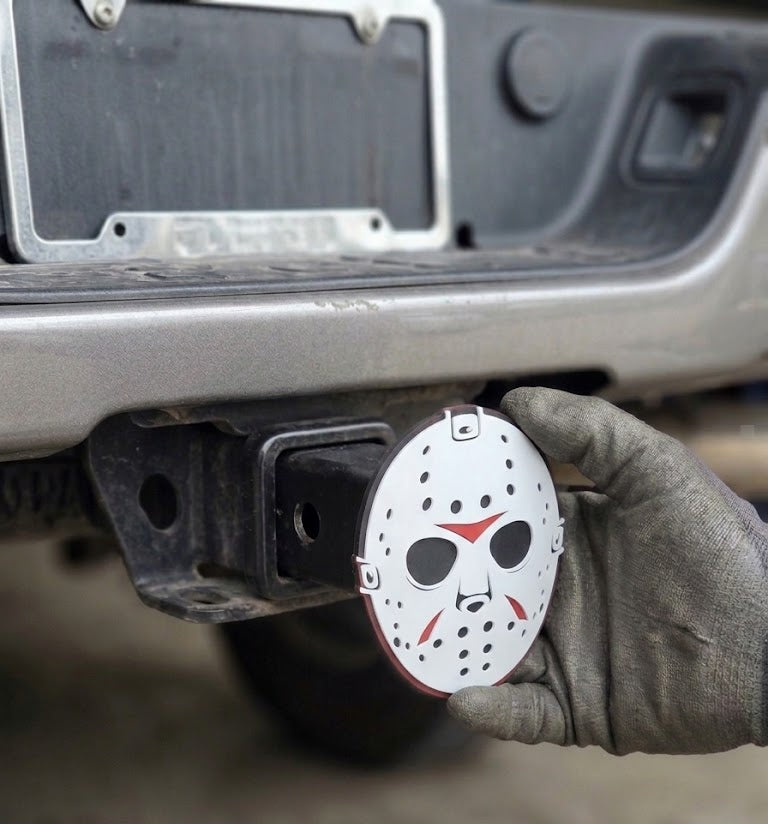 Hand holding a Hitch Cover with a Jason Voorhees mask design on a vehicle's hitch receiver.